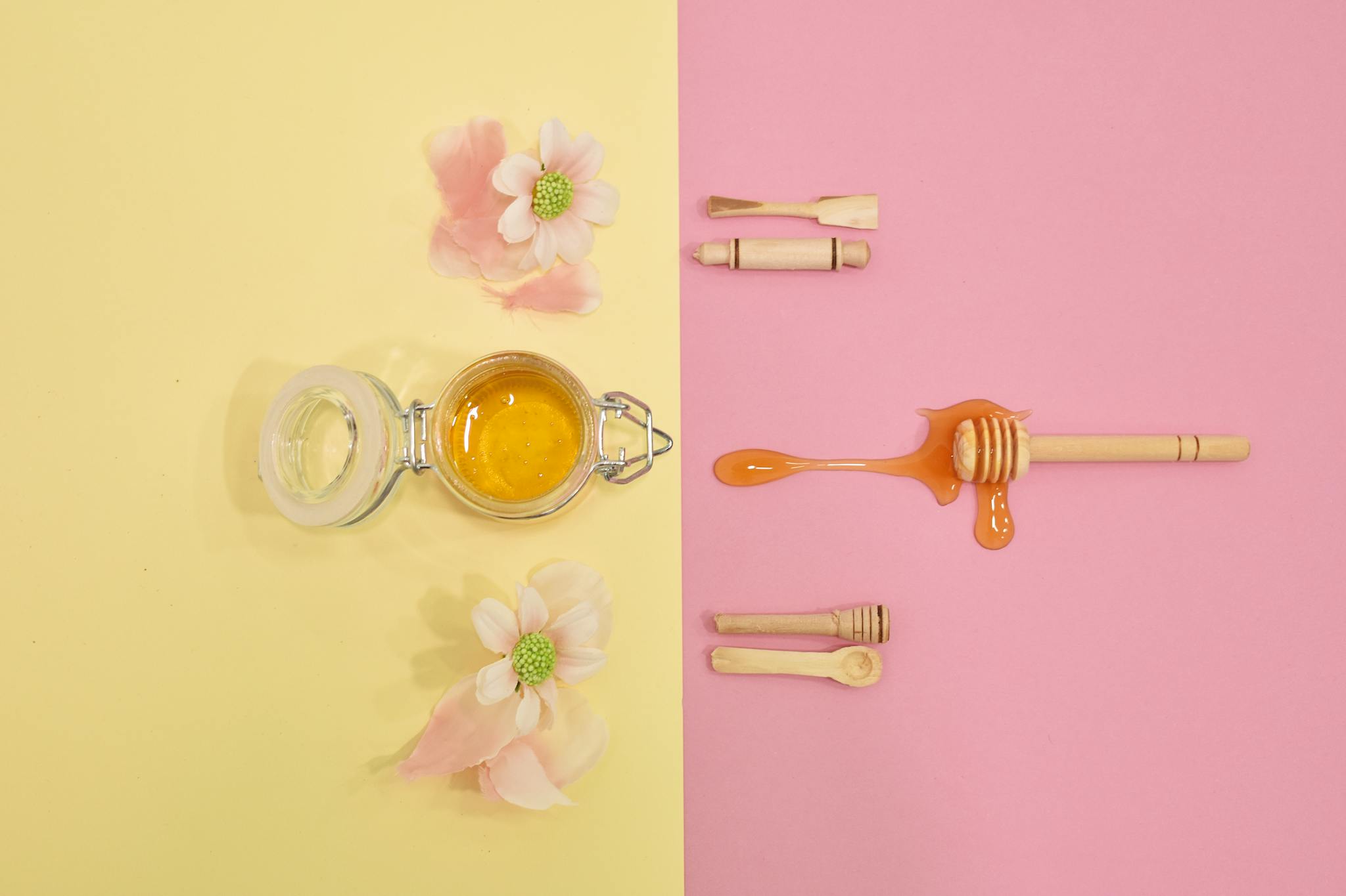 Top view of honey jar and wooden dipper with flowers on pastel backdrop.